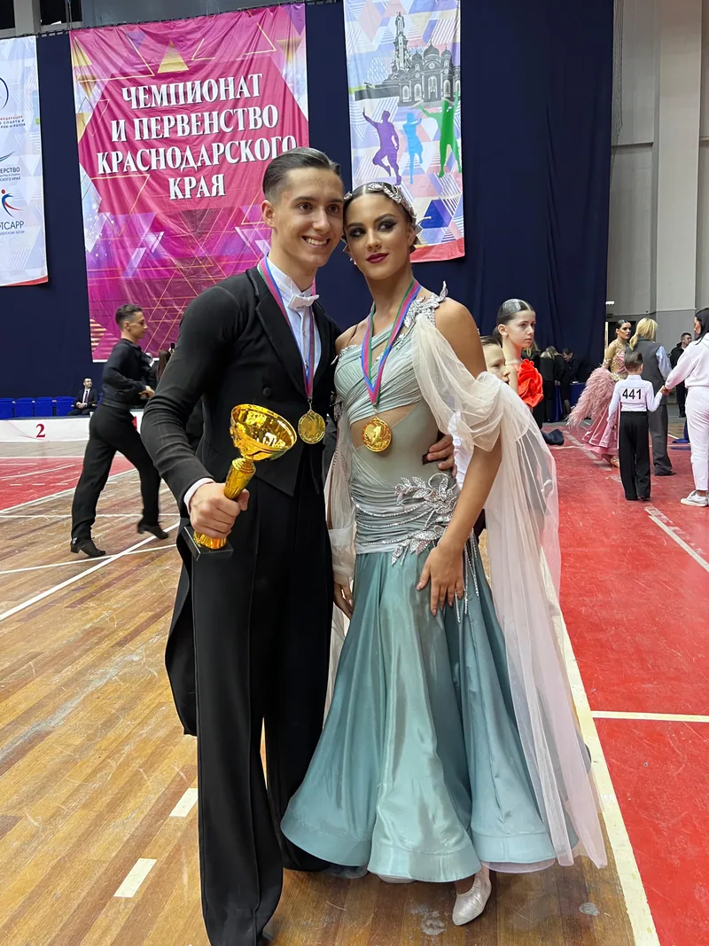 Пара в костюмах европейской программы с медалями и кубком у баннера чемпионата и первенства Краснодарского края — клуб «Дольче Вита» из Сочи